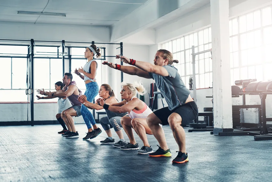A group of people in athletic wear squatting in unison with arms extended forward on a bright gym floor — gym and fitness studio insurance in New Jersey