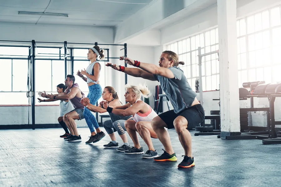 A group of people in athletic wear squatting in unison with arms extended forward on a bright gym floor — gym and fitness studio insurance in Iowa