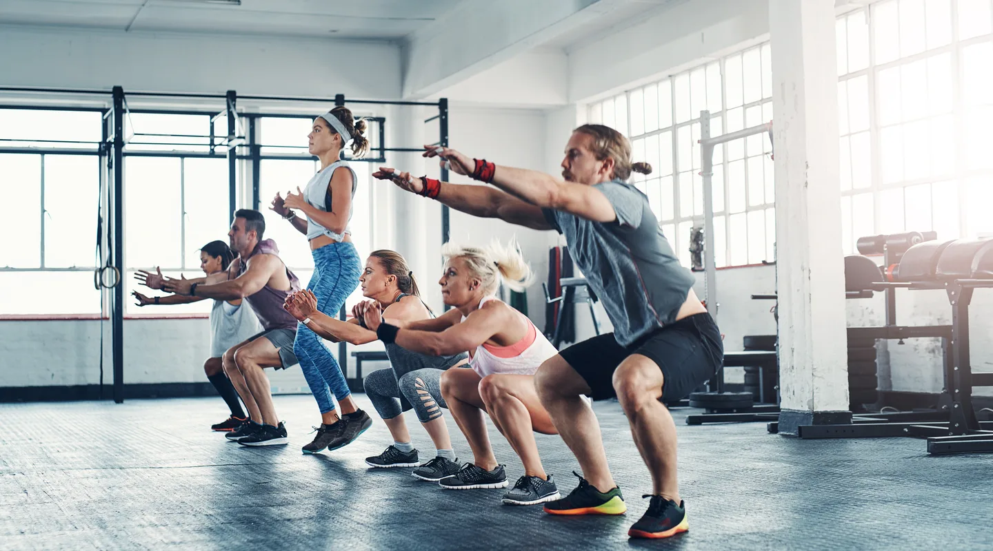 A group of people in athletic wear squatting in unison with arms extended forward on a bright gym floor