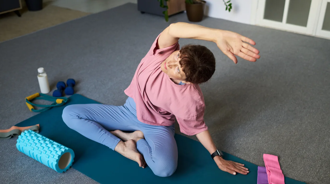A person seated on an exercise mat leaning into a side stretch, with a foam roller, water bottle and resistance bands nearby