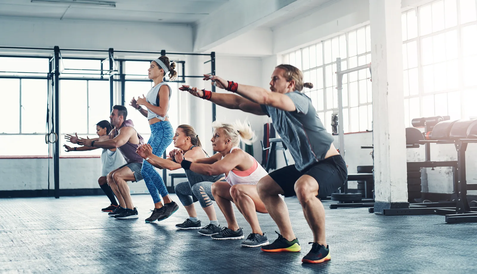 A group of people in athletic wear squatting in unison with arms extended forward on a bright gym floor
