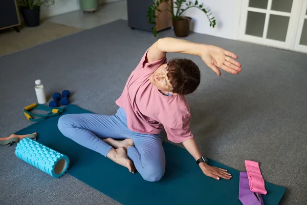 A person seated on an exercise mat leaning into a side stretch, with a foam roller, water bottle and resistance bands nearby