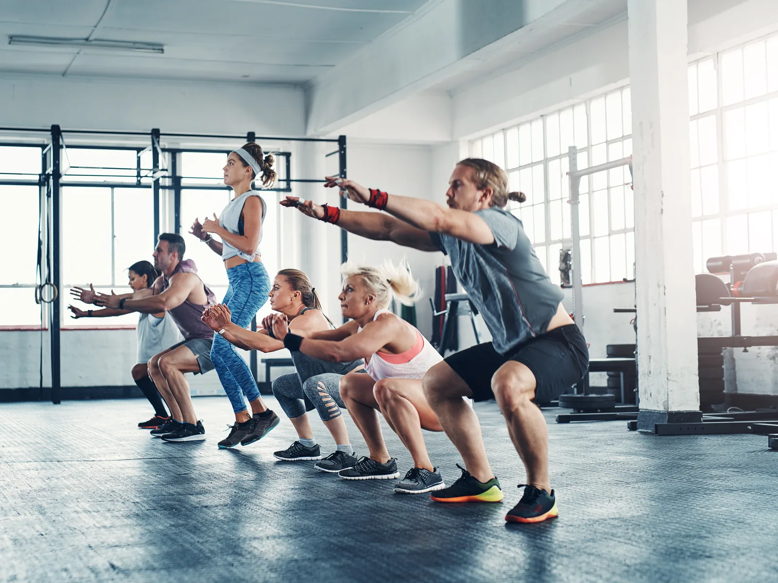 A group of people in athletic wear squatting in unison with arms extended forward on a bright gym floor