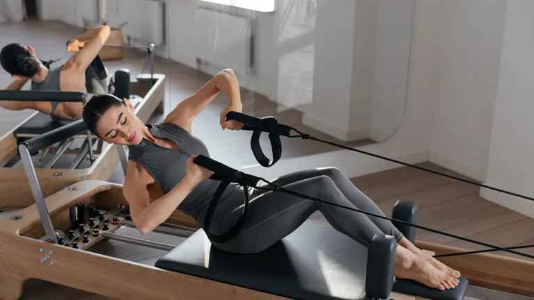 A person lying back on a reformer carriage and drawing the handles in a bright studio — gym and fitness studio insurance in Massachusetts