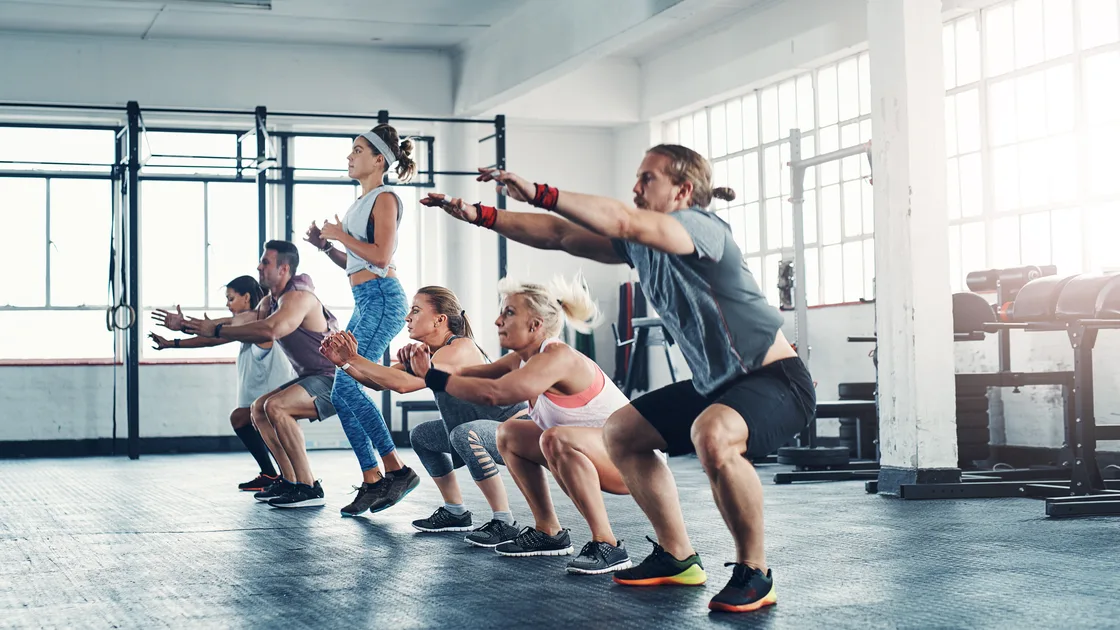 A group of people in athletic wear squatting in unison with arms extended forward on a bright gym floor — gym and fitness studio insurance in Mississippi
